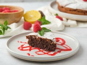 Horizontal shot of a slice of chocolate almond flour cake on top of raspberry puree on a white plate. A whole cake sits off to the right.