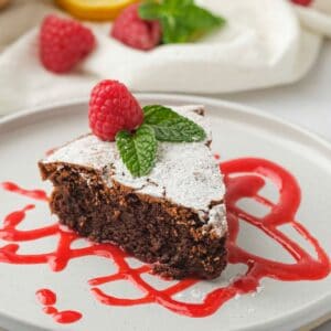 Horizontal shot of a slice of chocolate almond flour cake on top of raspberry puree on a white plate. A whole cake sits off to the right.