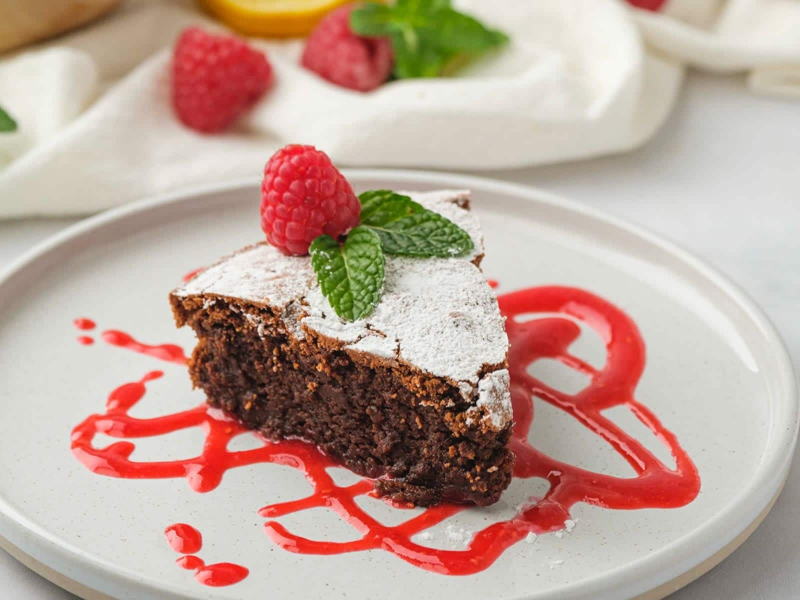 Horizontal shot of a slice of chocolate almond flour cake on top of raspberry puree on a white plate. A whole cake sits off to the right.