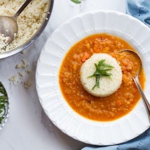 Red soup with a scoop of white rice and a green herb garnish in a white bowl next to a blue towel on a white marble background.