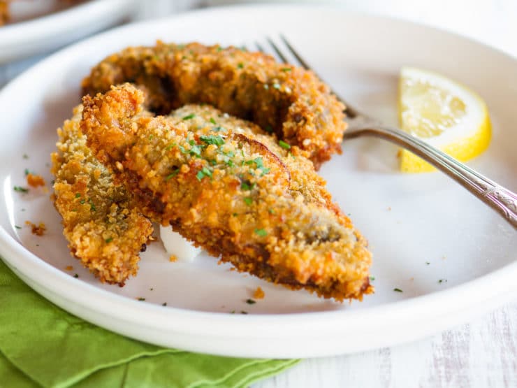 Vertical shot - Portobello Mushroom Schnitzel on a white plate with fork on side and a sliced lemon.