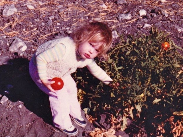 Tori Avey as a toddler in her grandfather\'s garden holding a tomato, leaning over a tomato plant.