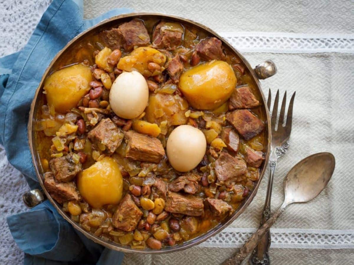 Top view of a Slow-cooked stew for Shabbat with potatoes, meat, and hard boiled eggs served on a fancy bowl with fancy spoon on fork on the left side