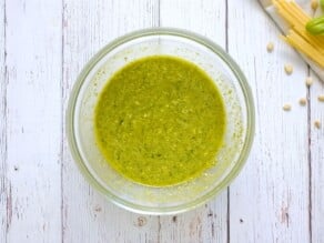 Pesto and pasta water sauce with olive oil mixed together in a glass bowl on a white wooden background, pine nuts and lemon on the side.