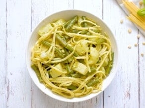 Overhead shot - bowl of trenette al pesto pasta with green beans and potatoes on a white wooden background, pine nuts and lemon on the side.