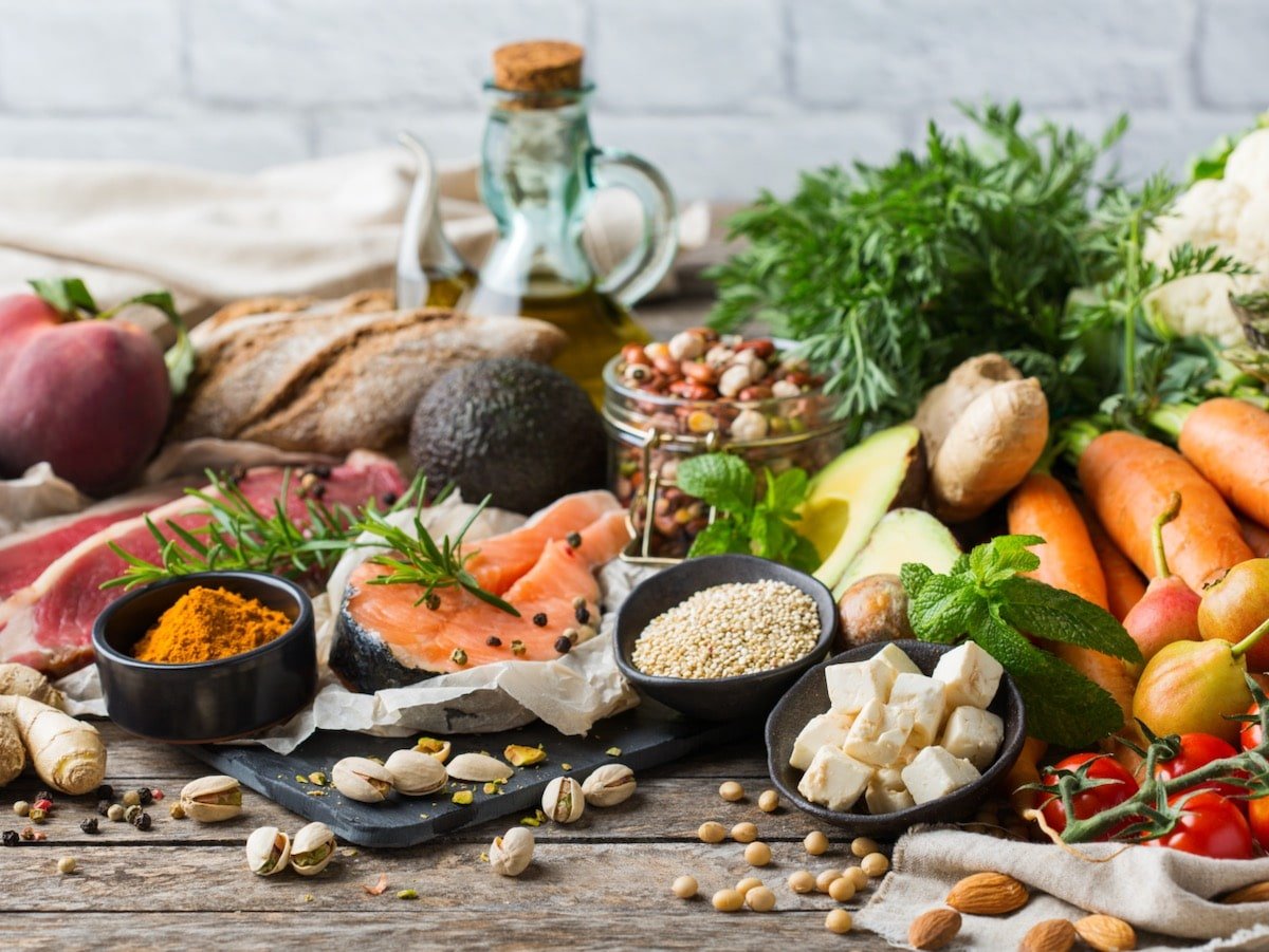 Wooden table covered with healthy Mediterranean ingredients - fresh salmon, vegetables, nuts, spices, fresh herbs, and a bottle of olive oil.