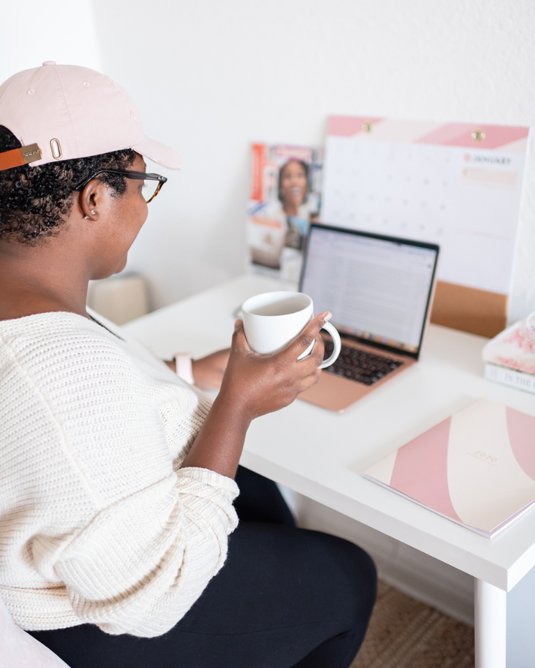 Black girl wearing athleisure wear sitting at her desk working on her business with a cup of coffee in her hand.
