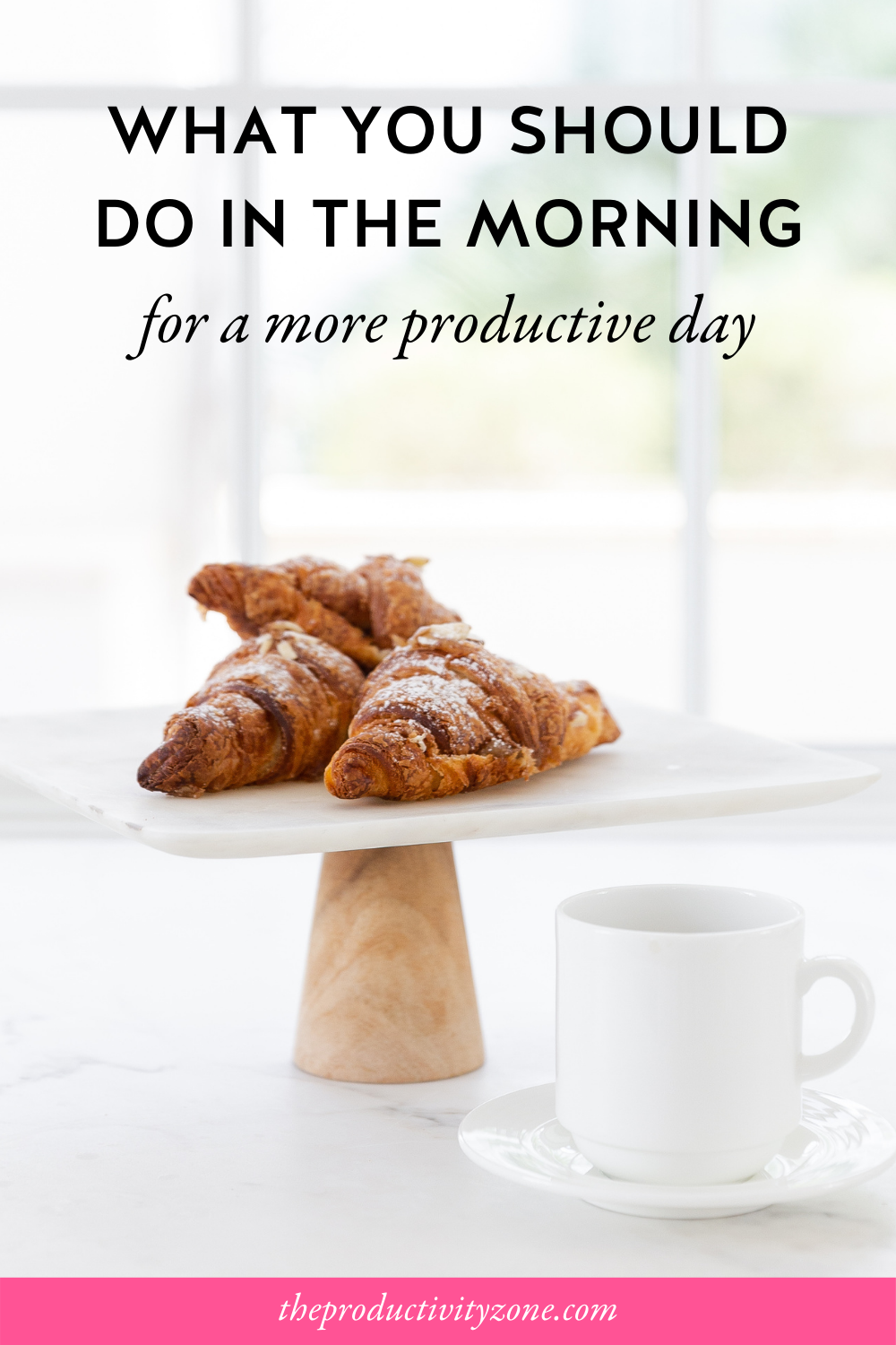 Chocolate croissants on a platter with a wooden pedestal beside a white coffee cup and saucer on a marble counter in front of a brightly lit window